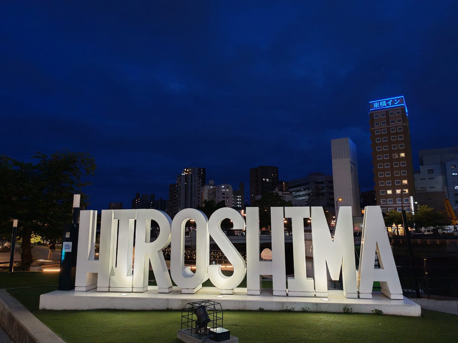 Grandes lettres lumineuses « HIROSHIMA » sur les berges de la rivière Motoyasu à Hiroshima au crépuscule, avec la skyline de la ville en arrière-plan.