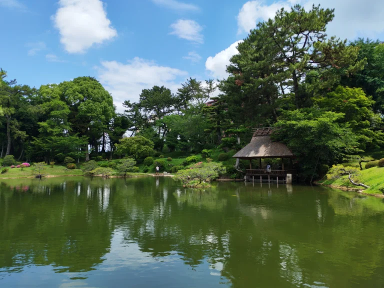 Vue du jardin Shukkei-en à Hiroshima avec son étang, un pavillon traditionnel japonais, des arbres luxuriants et leurs reflets dans l'eau sous un ciel bleu